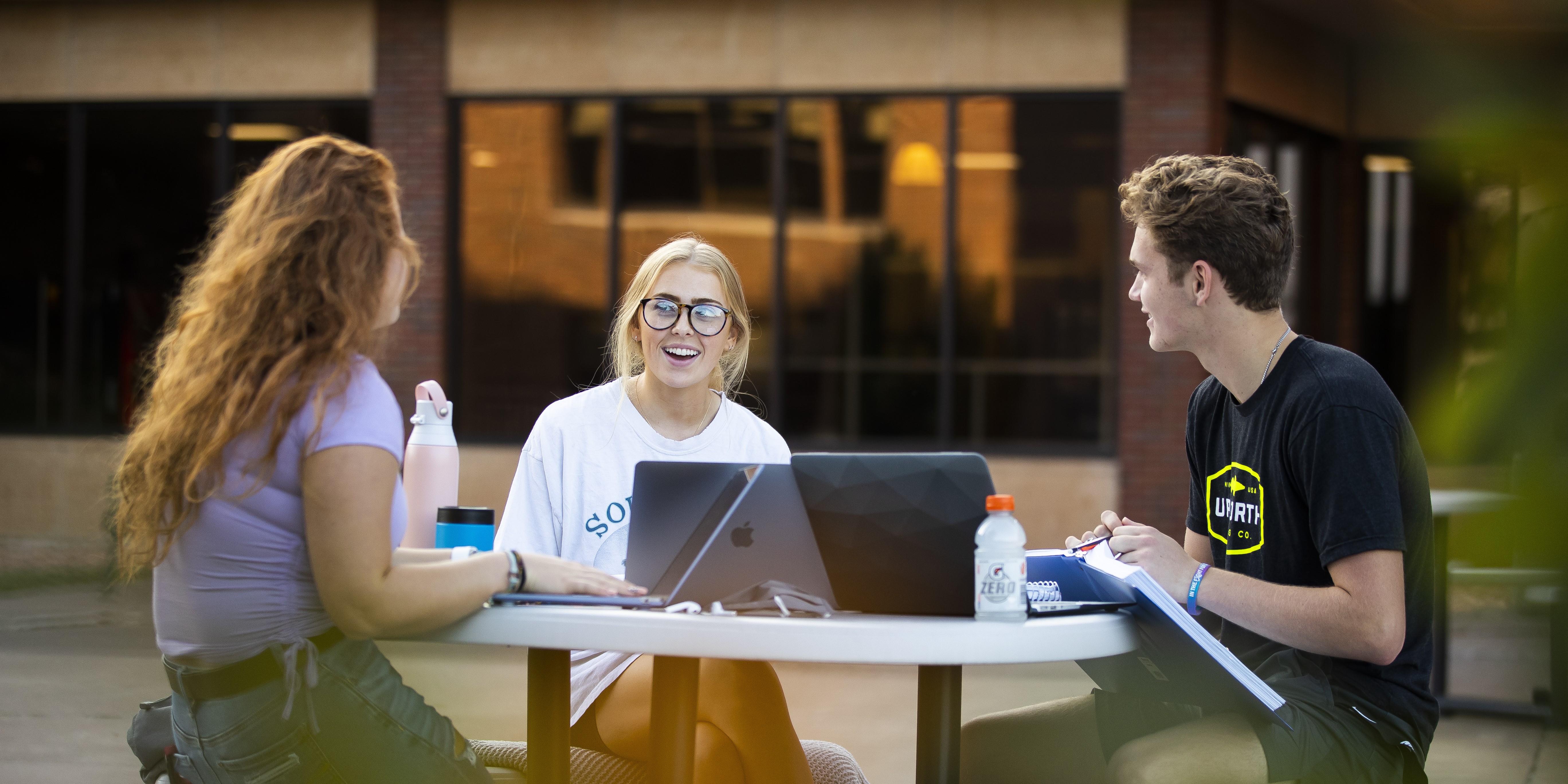 Three students study around a table outside with laptops