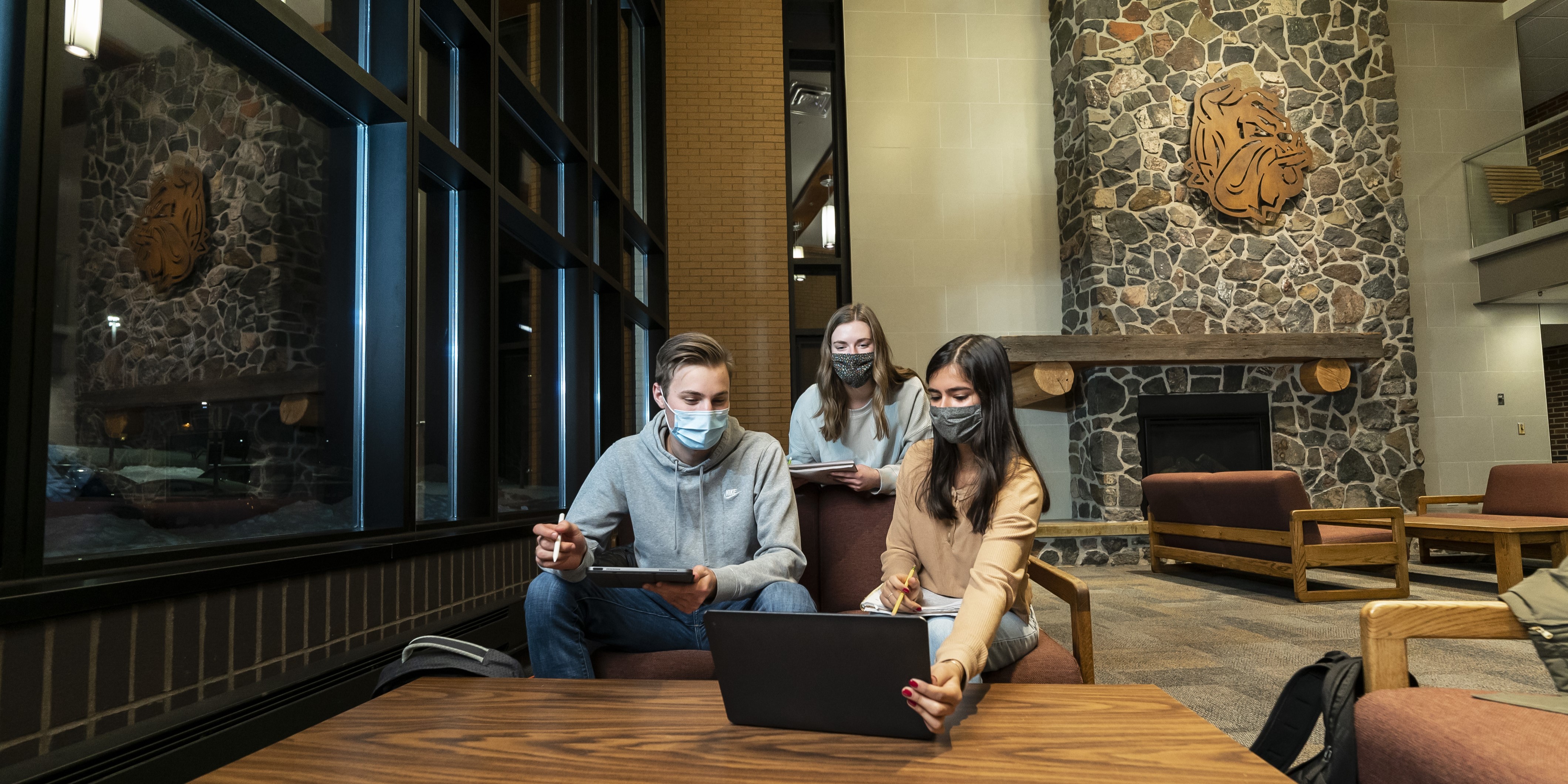 Three students, all wearing masks, study together with a laptop, tablet, and notebook